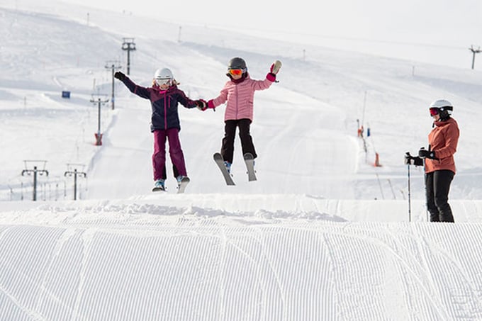 Two children jump on slalom skis in the ski resort while mother watches from the side